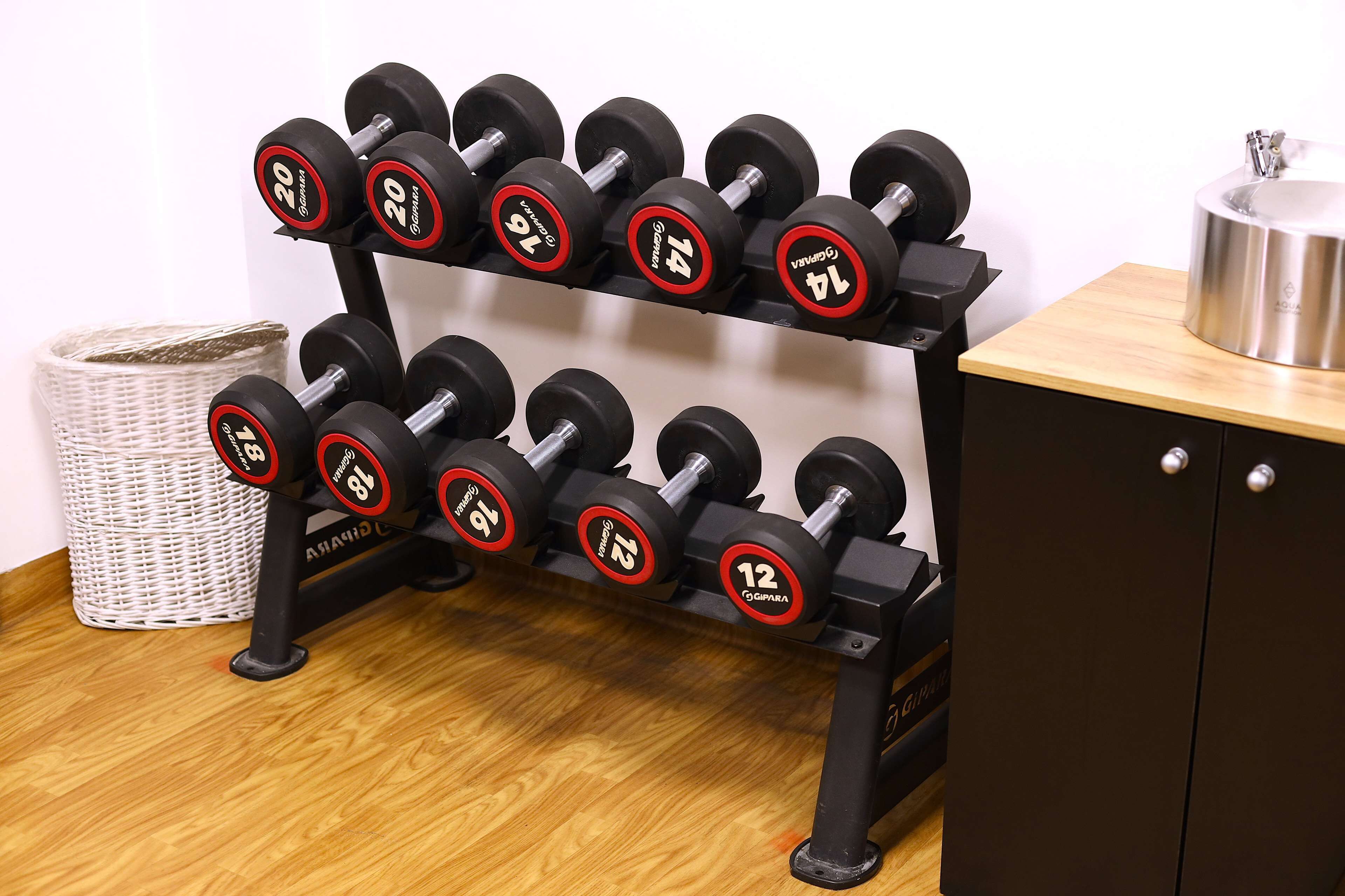 Een zwarte dumbbellrek met gewichten variërend van 12 tot 20 kilogram, een rieten mand en een kast met een roestvrijstalen bak erop.