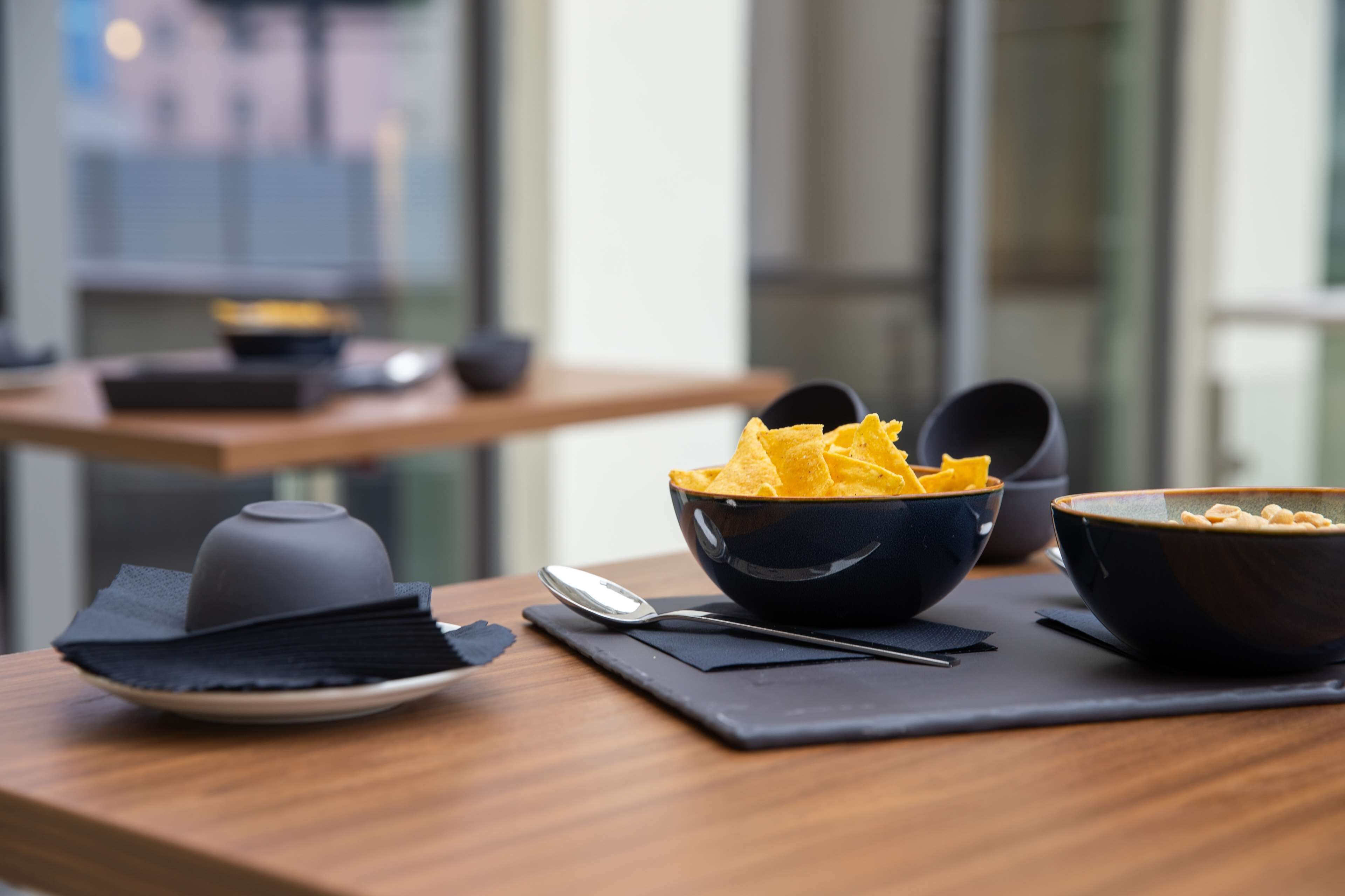 A dining table with bowls of chips and a teapot, set on a wooden table with a black placemat.