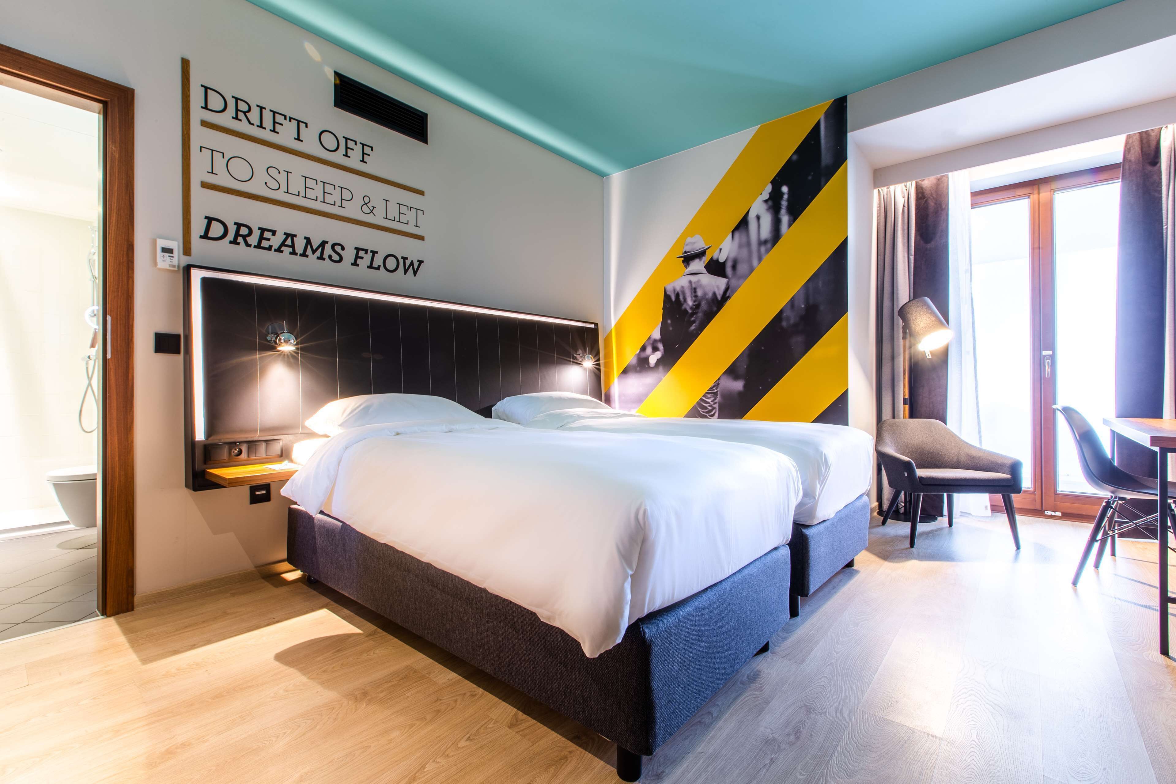 A modern hotel room with a large bed, a desk, and a chair. There is a wall with a motivational quote and a large poster.