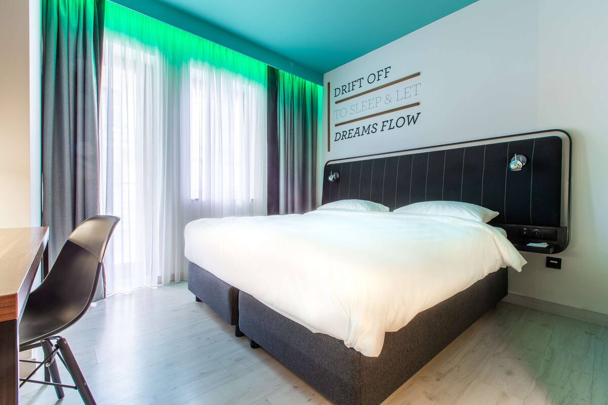 A modern hotel room with a large bed, a desk, and a view of the city through sheer curtains.