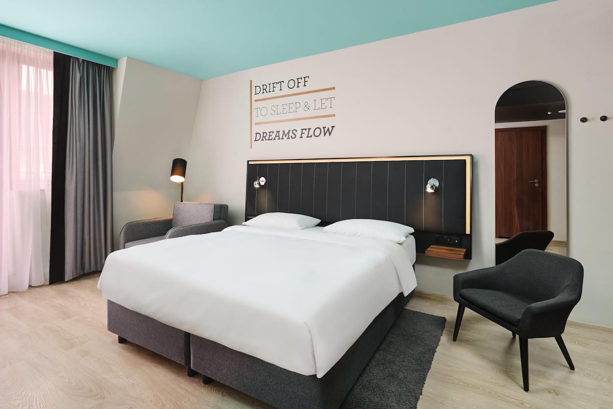 A modern hotel room with a large bed, a bedside table, a chair, and a wall-mounted TV.