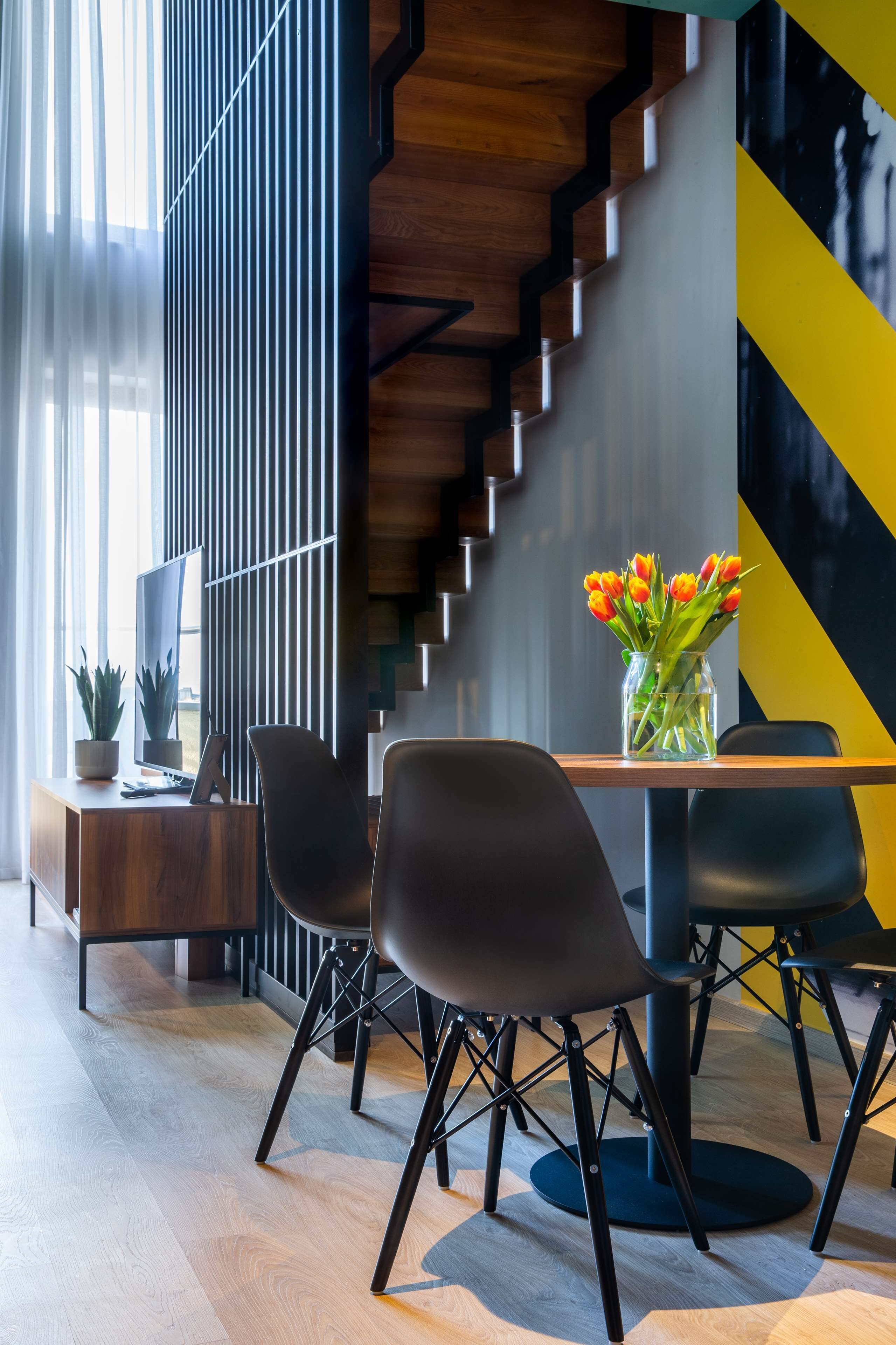 A modern dining area with a wooden table, black chairs, and a vase of orange tulips.
