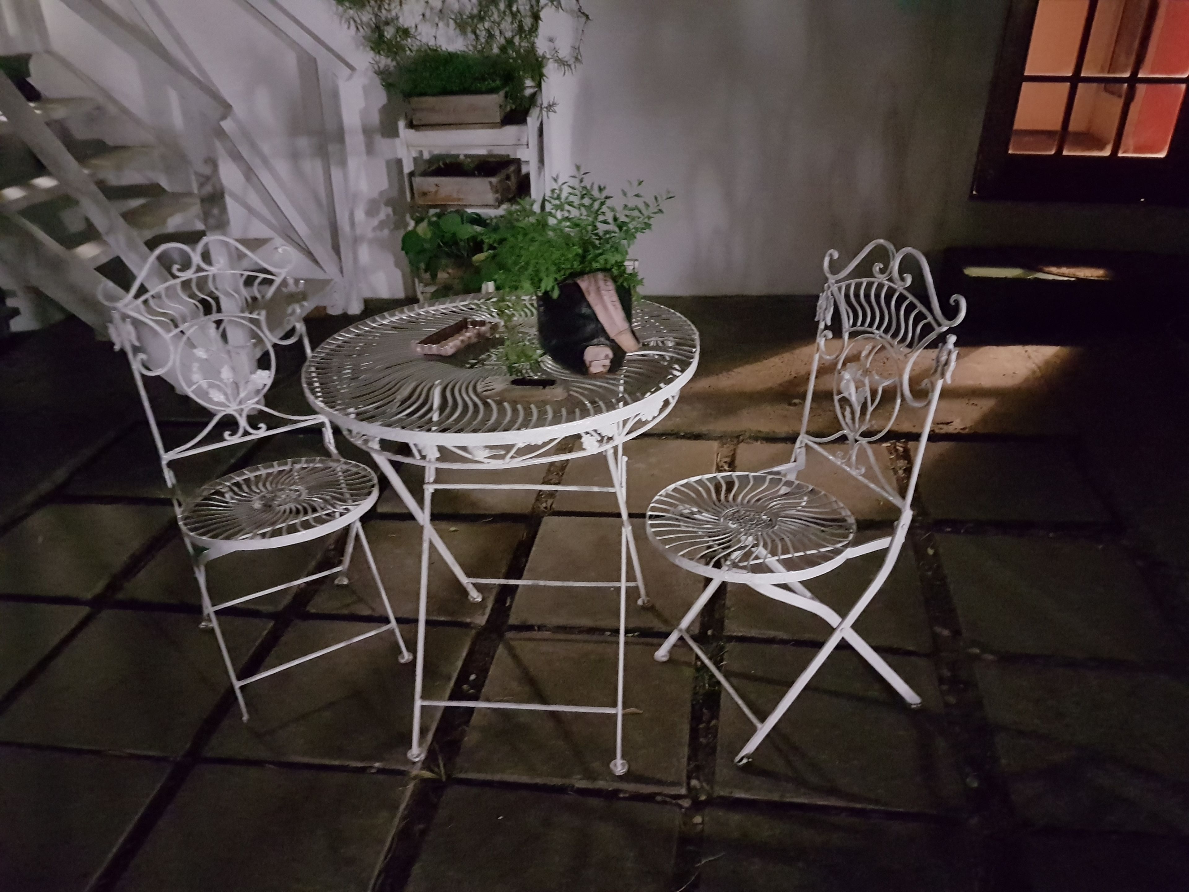 A round metal table with four chairs, surrounded by potted plants and a staircase.