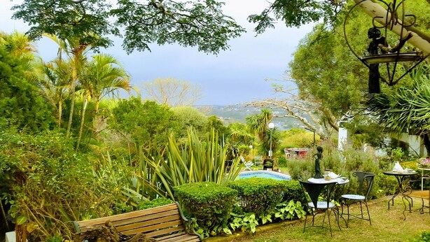 A lush garden with a wooden bench, metal tables, and chairs, surrounded by various plants and trees. - On-the-Summit Lodge & Conference Venue (East London)