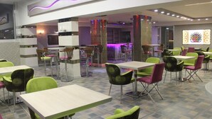 Un restaurant moderne avec des chaises vertes et roses, des tables blanches et un éclairage d’ambiance violet.