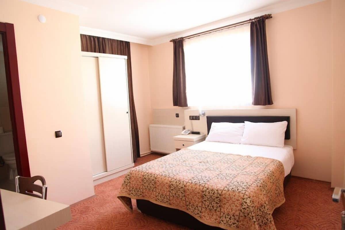 A hotel room with a bed, a bedside table, a chair, a window with curtains, and a door.