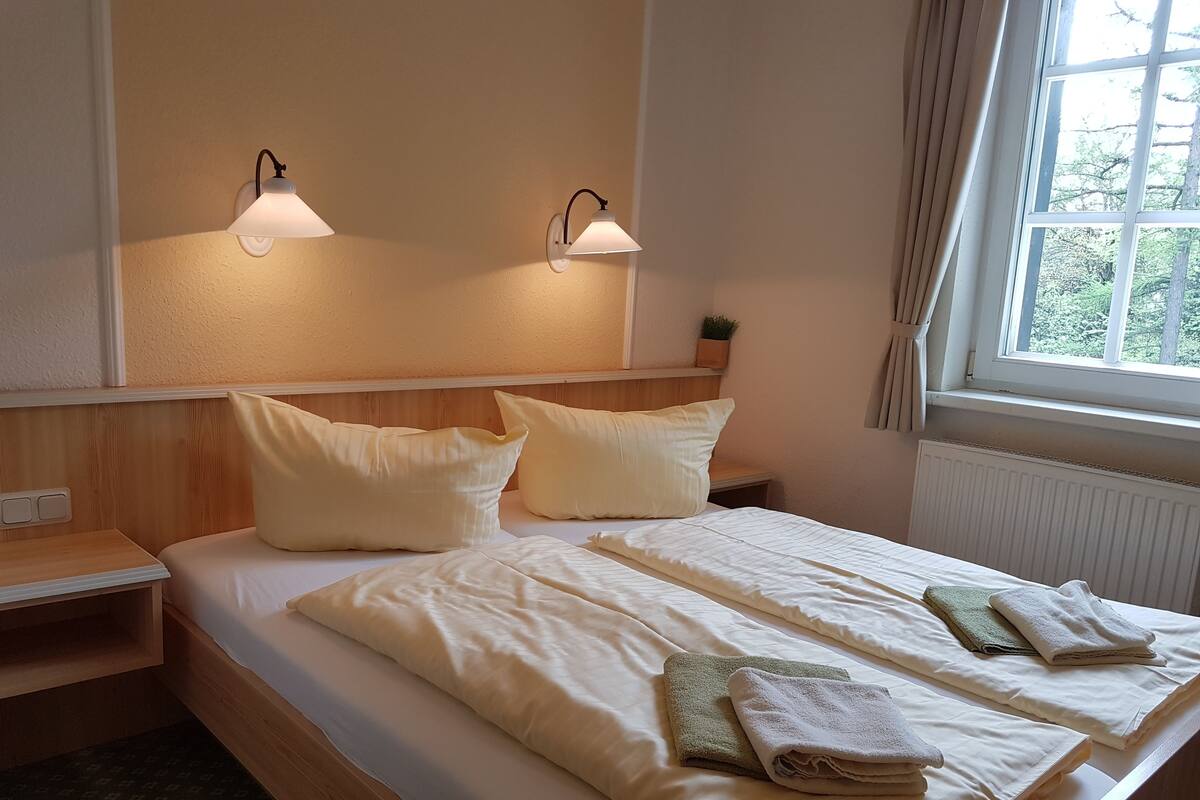 Habitación de hotel con cama, dos mesitas de noche, dos lámparas de pared, ventana con cortinas y un radiador.