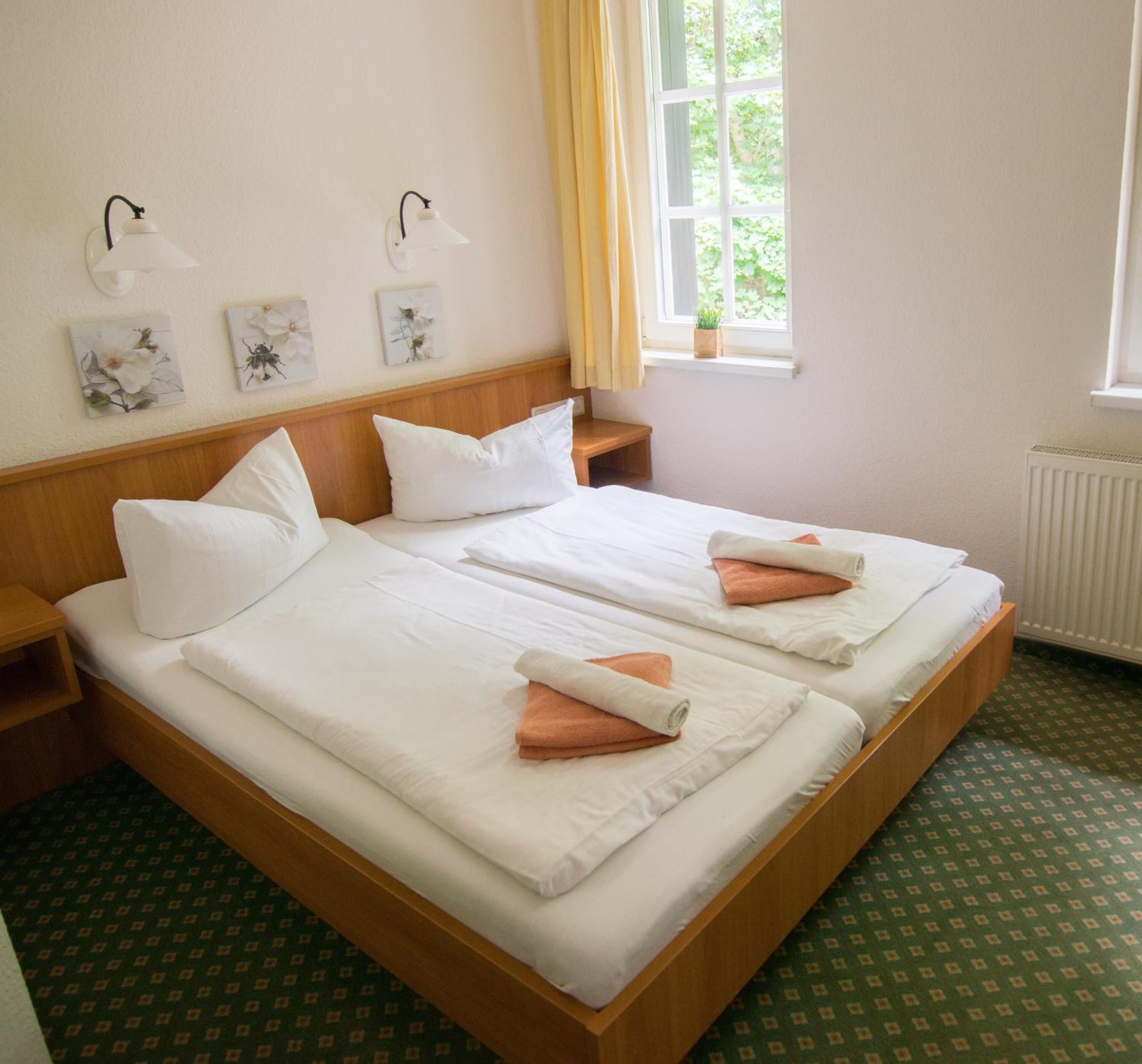 Cama doble con ropa de cama blanca y toallas naranjas, cabecera de madera, mesitas de noche, lámparas de pared y una ventana con cortinas.