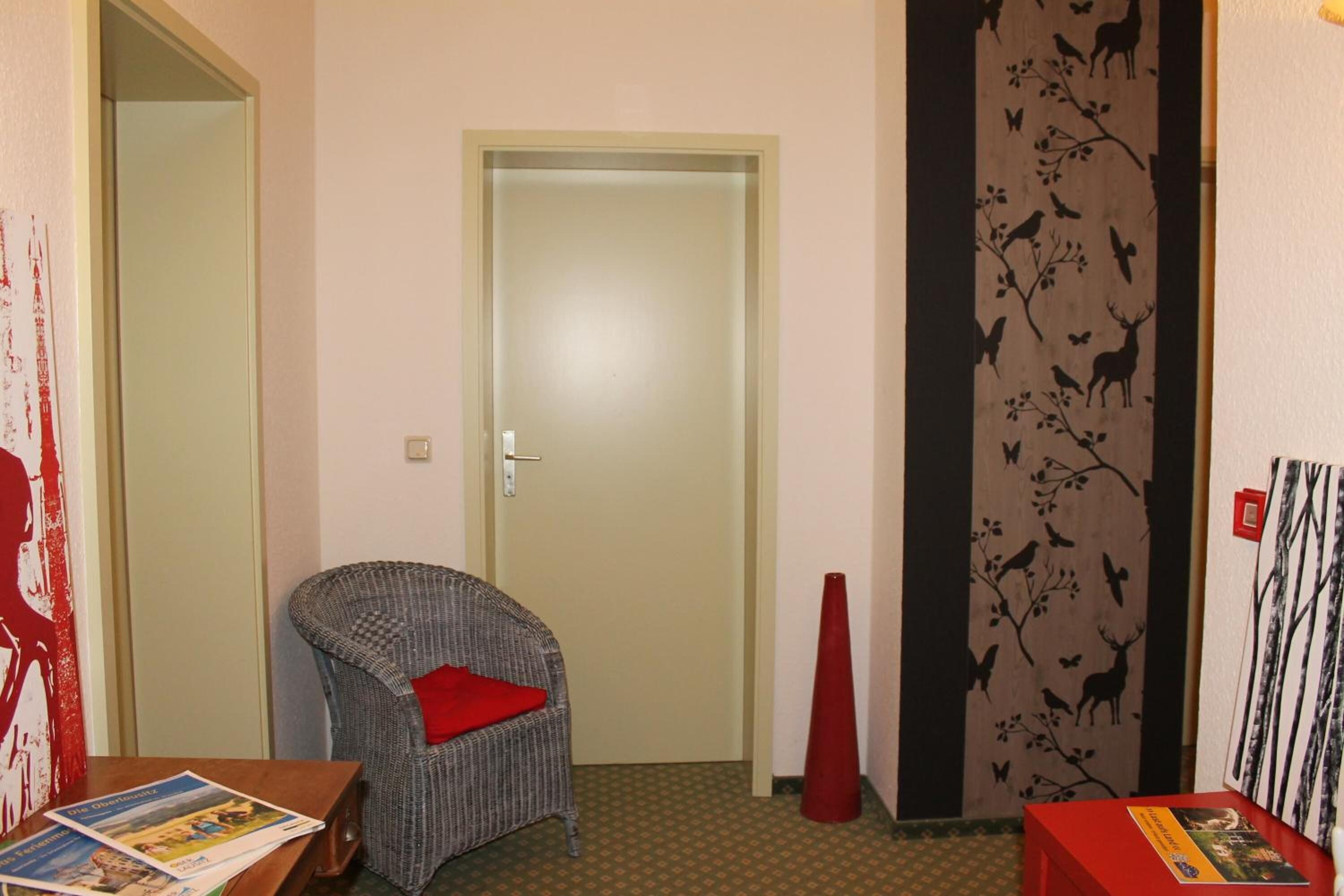 A room with a closed door, a wicker chair with a red cushion, a small table with magazines, and a red vase.