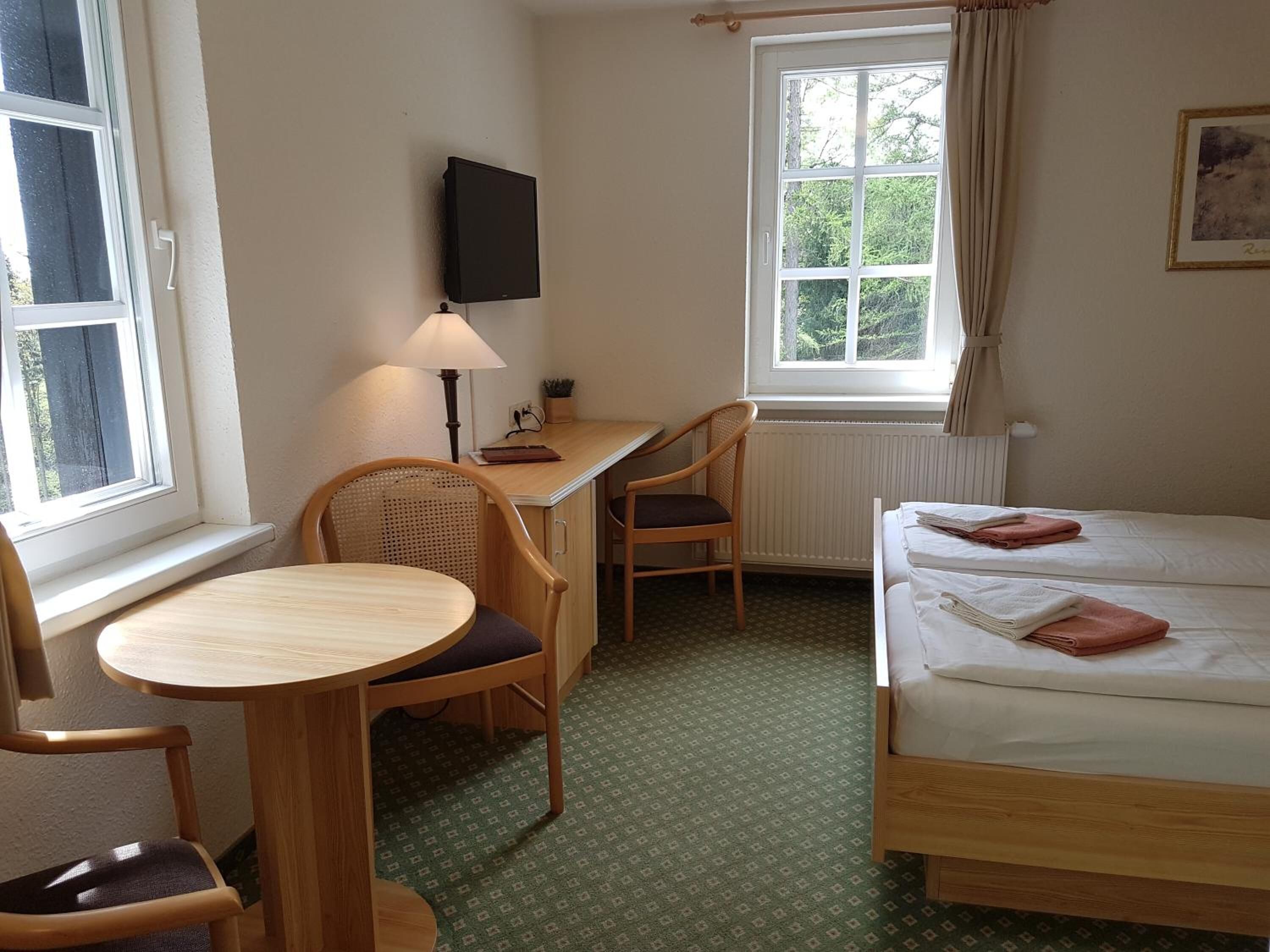 A small, well-lit room with a single bed, a desk with a chair, a table, and a TV mounted on the wall.