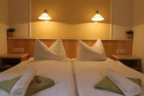 Une chambre d’hôtel avec deux lits, chacun muni d’un oreiller et d’une serviette enroulée posée sur une serviette verte. Des lampes murales sont fixées au-dessus des lits, et une petite plante orne la table de chevet.