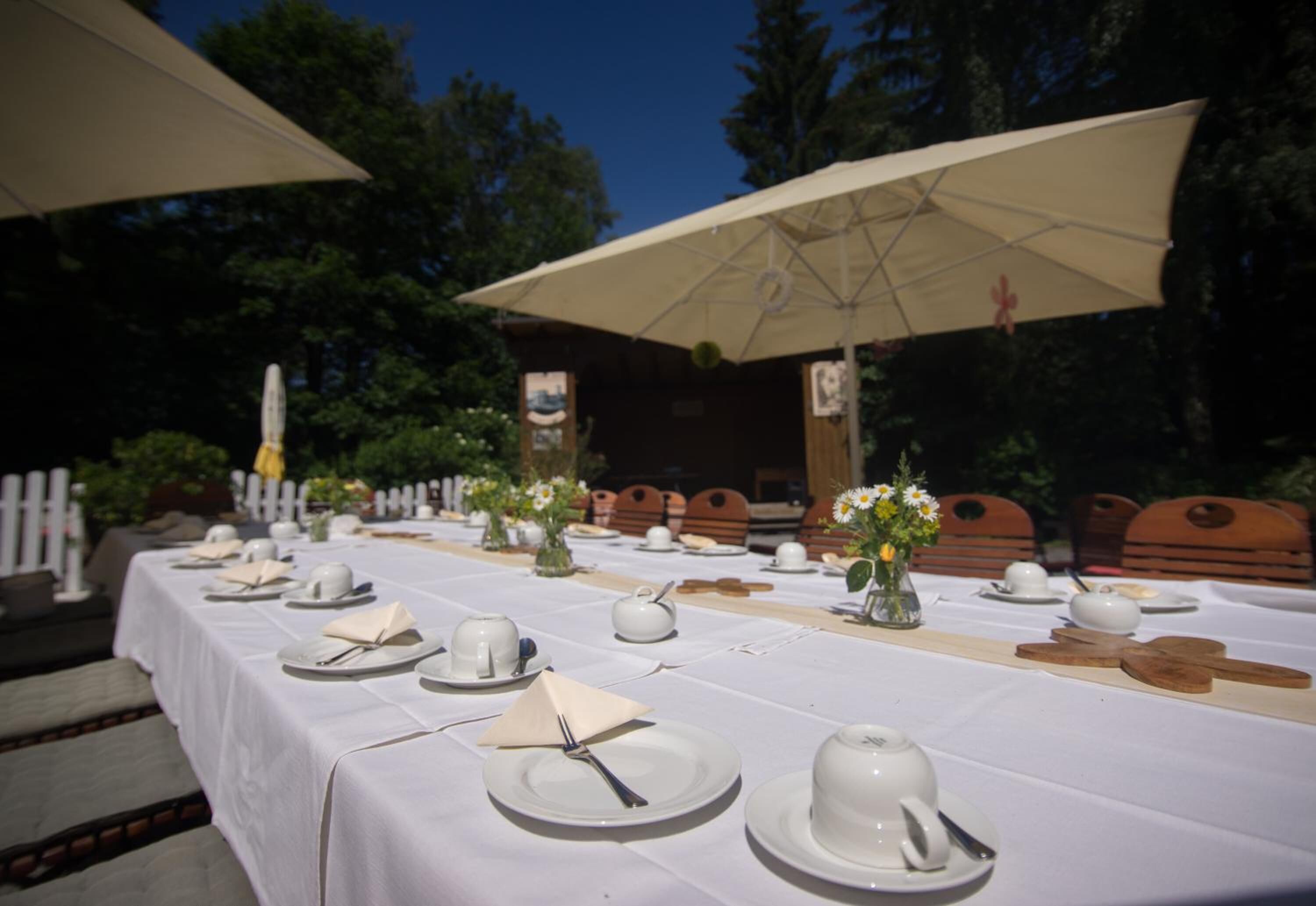 A table set for a meal outdoors with white tablecloths, plates, cups, and cutlery.