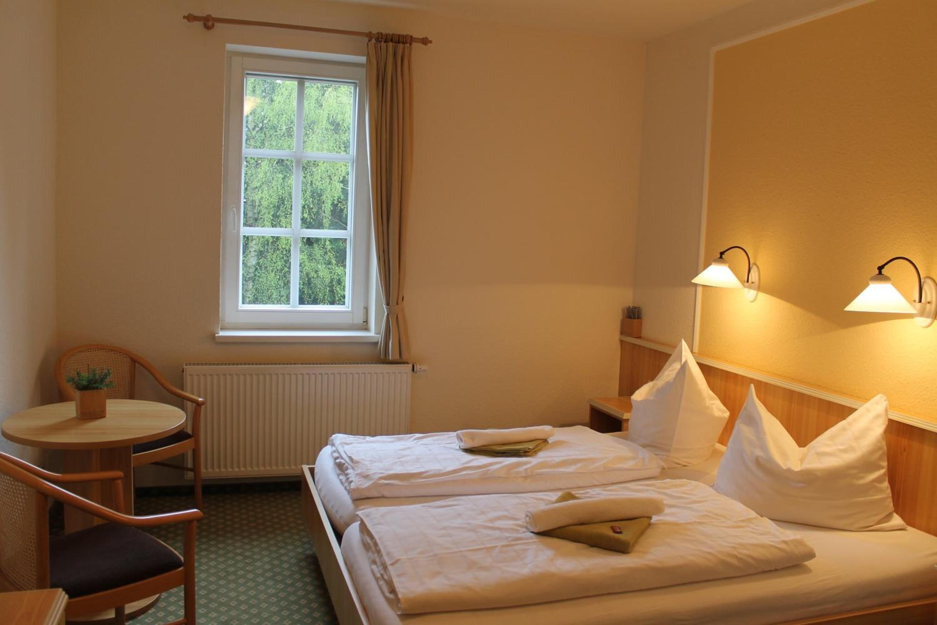 Een hotelkamer met twee bedden, een tafeltje met een stoel, een raam met gordijnen en wandlampen.