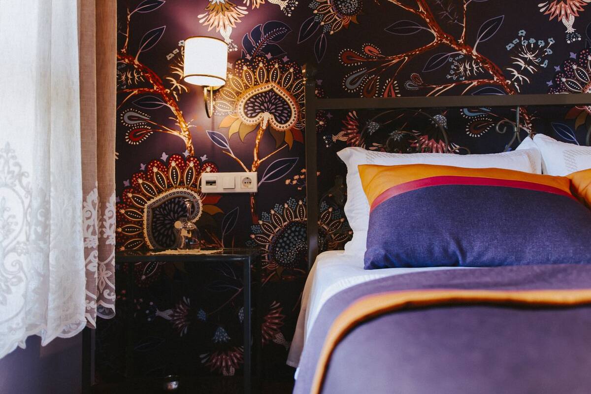 Un dormitorio con papel tapiz estampado, una lámpara de noche, una cama con muchos almohadones y una mesita de noche con una pequeña estatua.