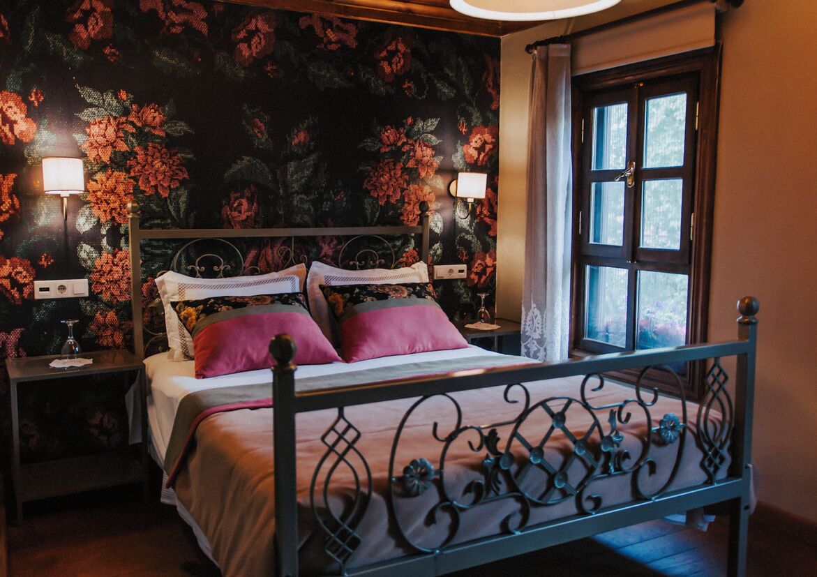 Un dormitorio con papel tapiz floral, una cama con ropa de cama rosa y blanca, dos mesitas de noche con lámparas y una ventana con cortinas.