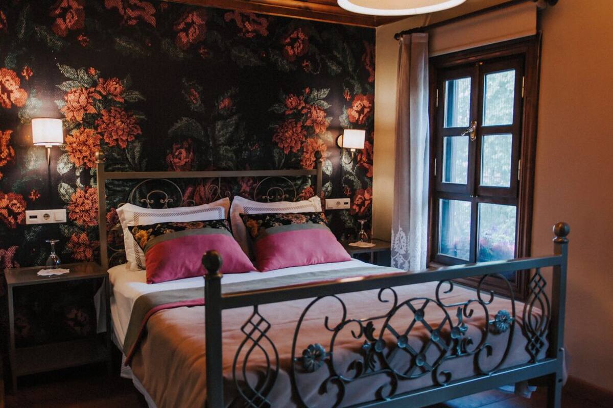 Un dormitorio con papel tapiz floral, una cama con estructura metálica, dos mesitas de noche con lámparas y una ventana con cortinas.