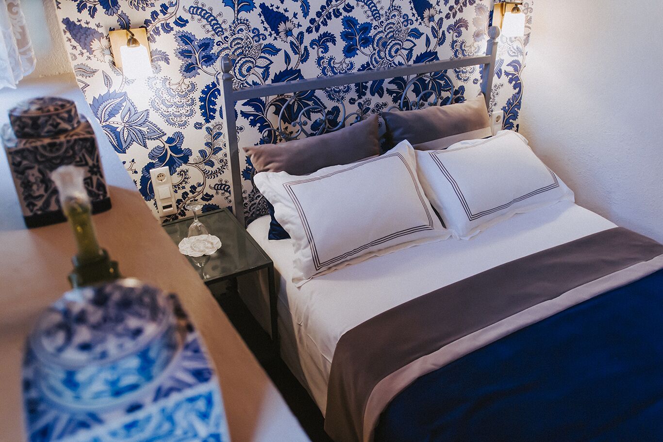Una cama bien hecha con ropa de cama blanca y marrón, un cabecero de metal y una mesita de noche con un vaso y un adorno.