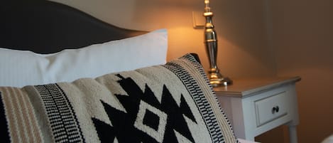 Una cama con una almohada estampada, una lámpara de noche y una mesita de noche pequeña blanca con un cajón.