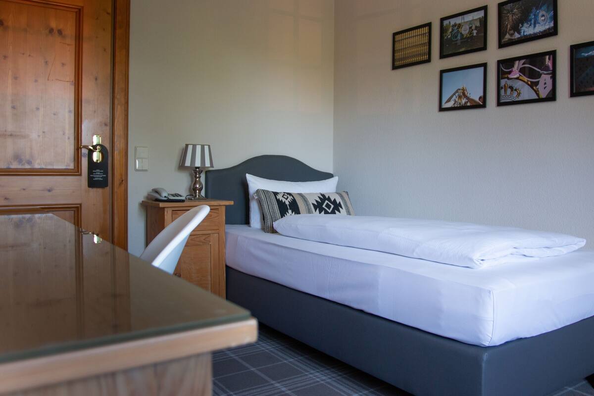 A hotel room with a bed, a desk, a chair, a lamp, and a door.