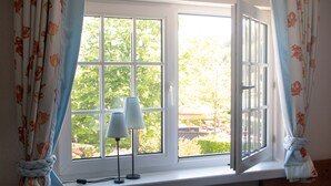 Ein Fenster mit Blick auf einen Garten und eine Terrasse, Vorhänge zurückgebunden, und eine Lampe im Fensterbrett.