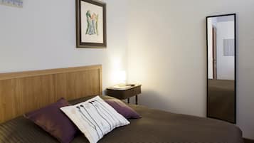 Un letto rifatto con cura, una testiera, due cuscini, un comodino con una lampada e un quadro incorniciato appeso al muro.