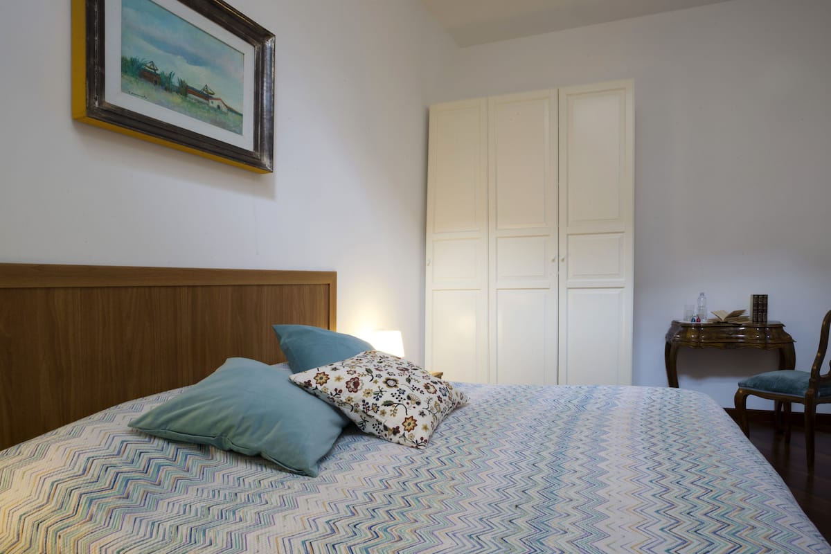 Un dormitorio con una cama, almohadas, una mesita de noche, una silla, un armario y un cuadro en la pared.
