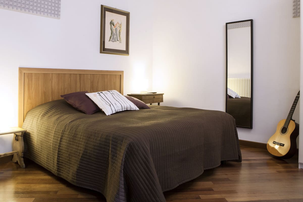 Un dormitorio ordenado con un cabecero de madera, una cama con un almohadón a rayas, una mesita de noche con una lámpara, una guitarra y un espejo grande.