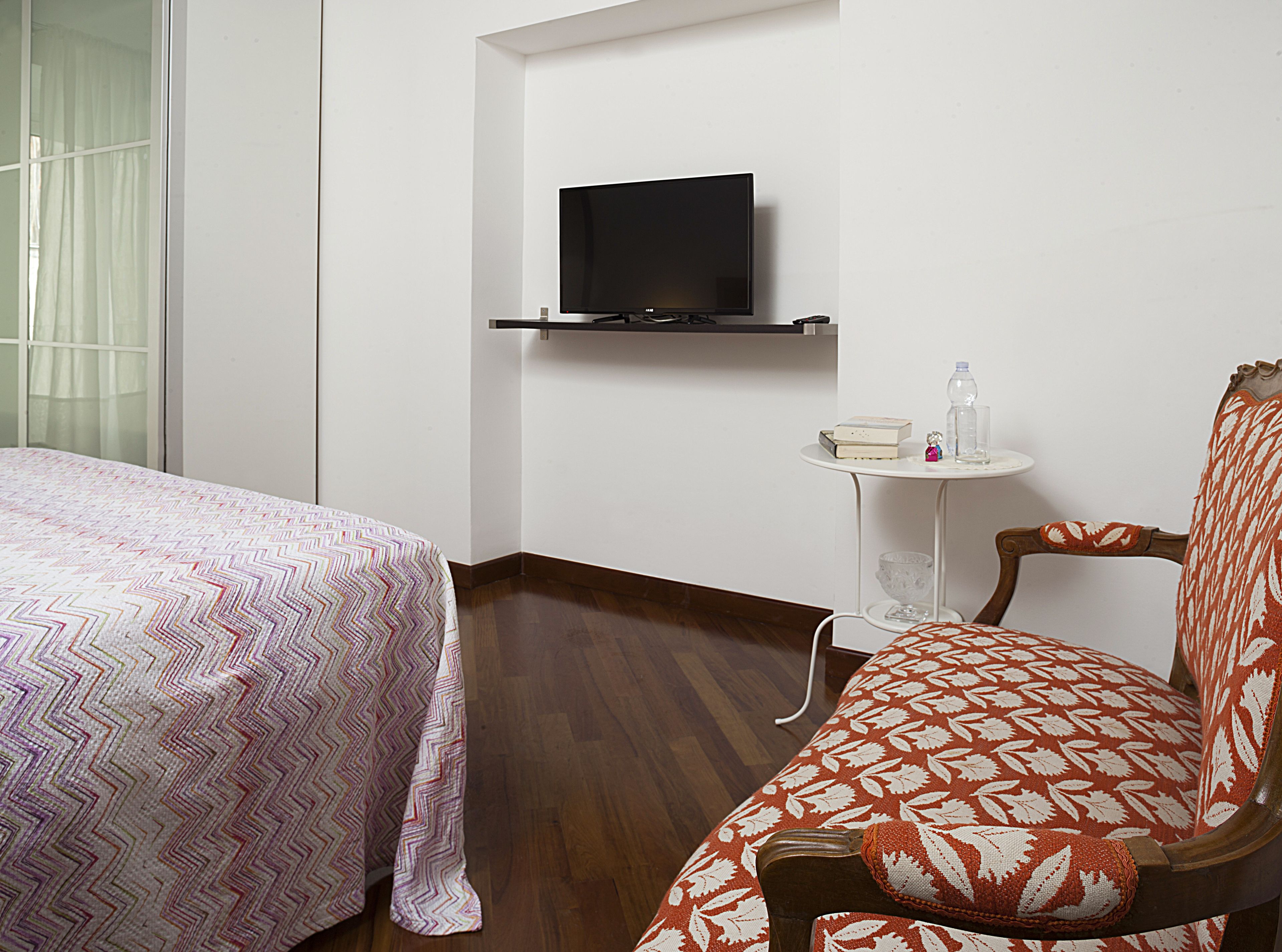 A hotel room with a bed, a chair, a small table with a bottle and a glass, and a TV mounted on the wall.
