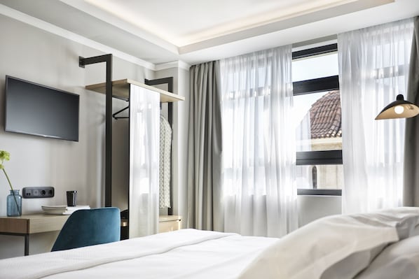 A hotel room with a bed, a desk with a chair, a TV mounted on the wall, and a window with curtains.
