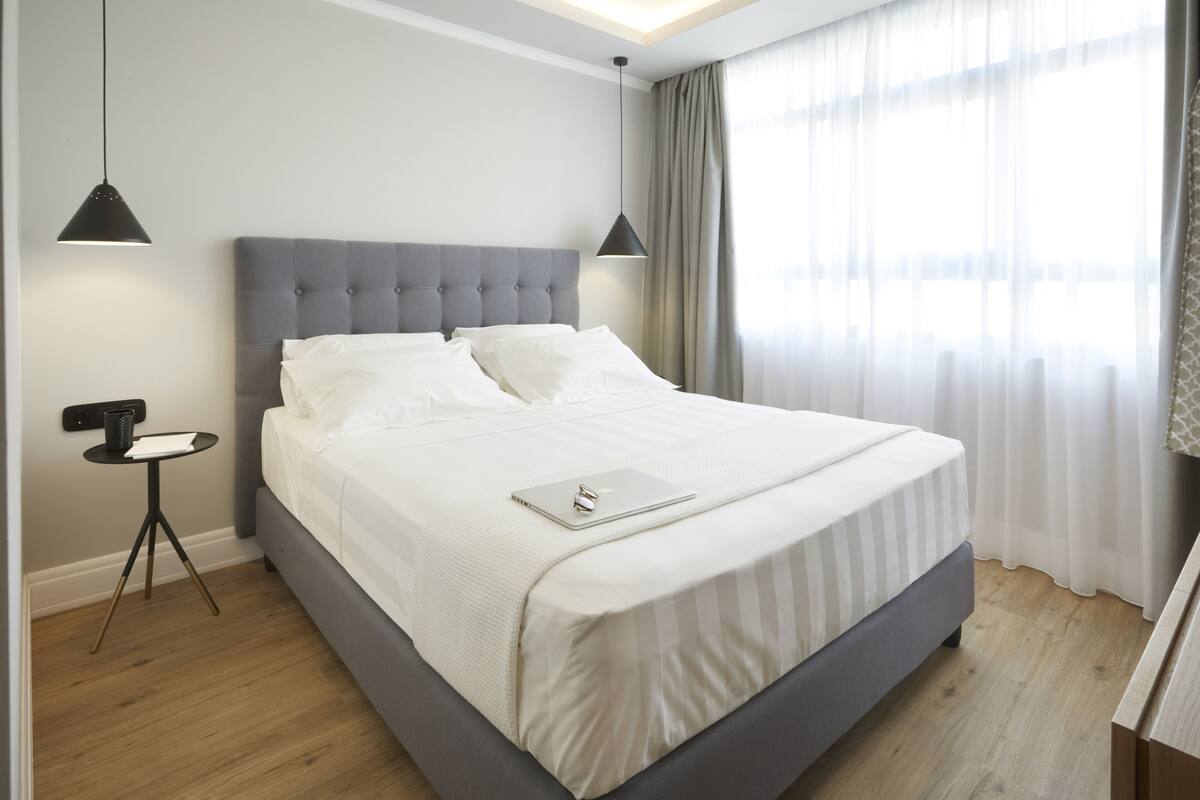 Une chambre à coucher moderne avec un grand lit, deux lampes suspendues, une petite table de chevet et une vue sur l’extérieur à travers des rideaux légers.