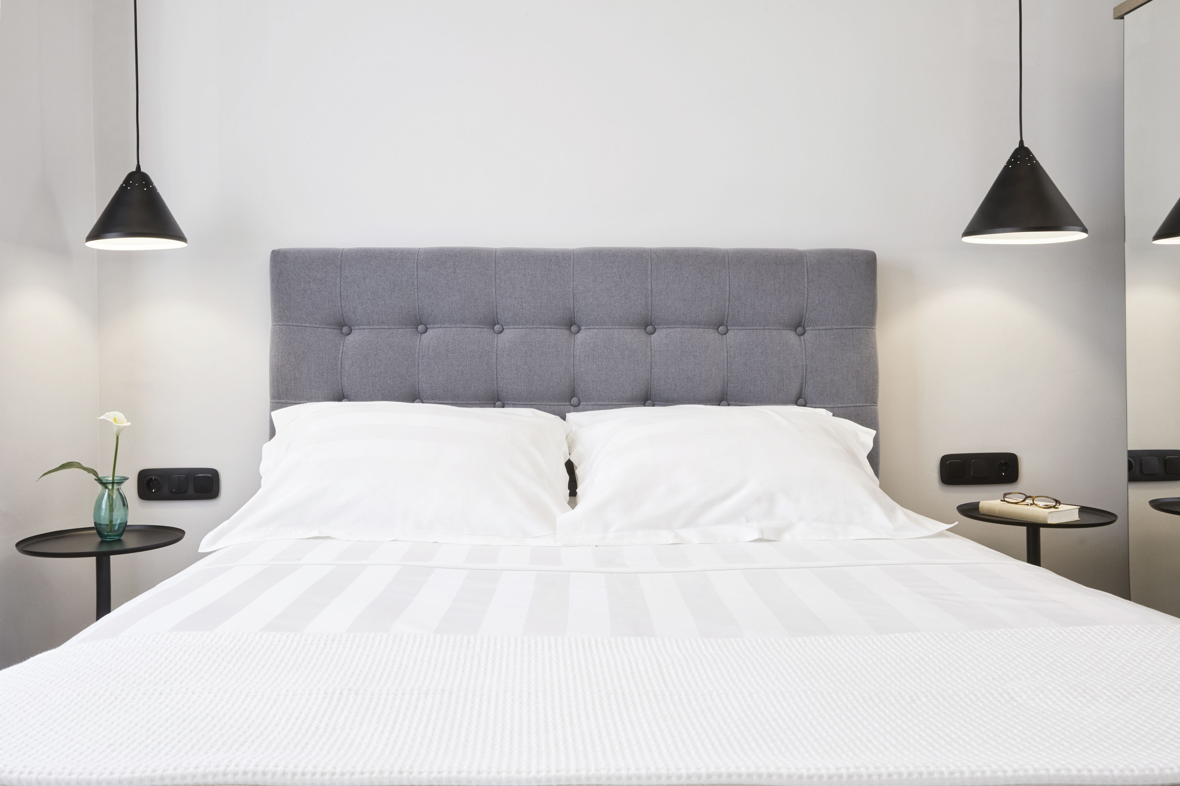 Una cama bien hecha con un cabecero tapizado gris, almohadas blancas y una colcha blanca. Hay dos mesitas de noche, cada una con una lámpara negra y un pequeño jarrón con una flor blanca.