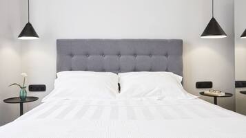 Una cama bien hecha con un cabecero tapizado gris, almohadas blancas y una colcha blanca. Hay dos mesitas de noche, cada una con una lámpara negra y un pequeño jarrón con una flor blanca.