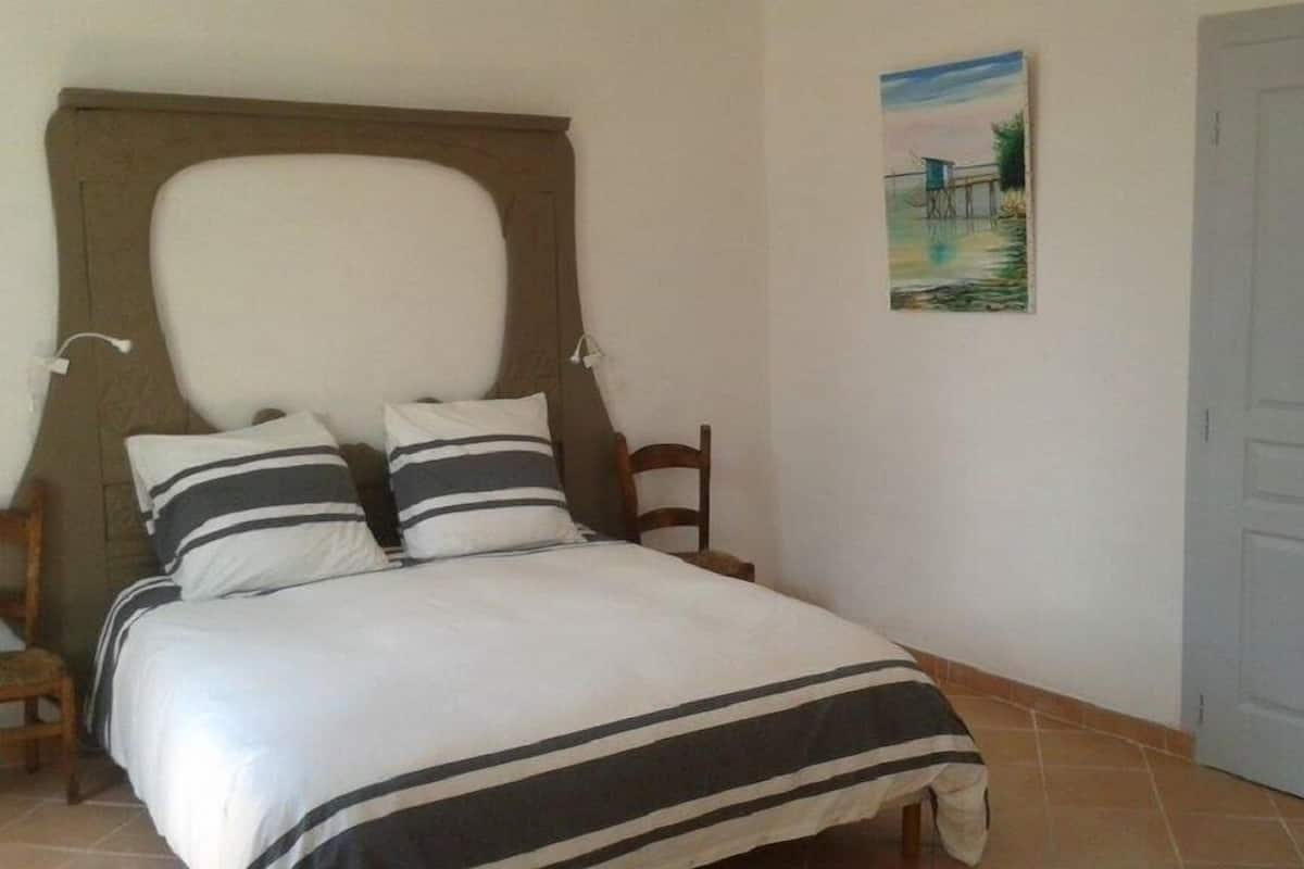 A bedroom with a bed, two pillows, a wooden chair, a painting on the wall, and a door.