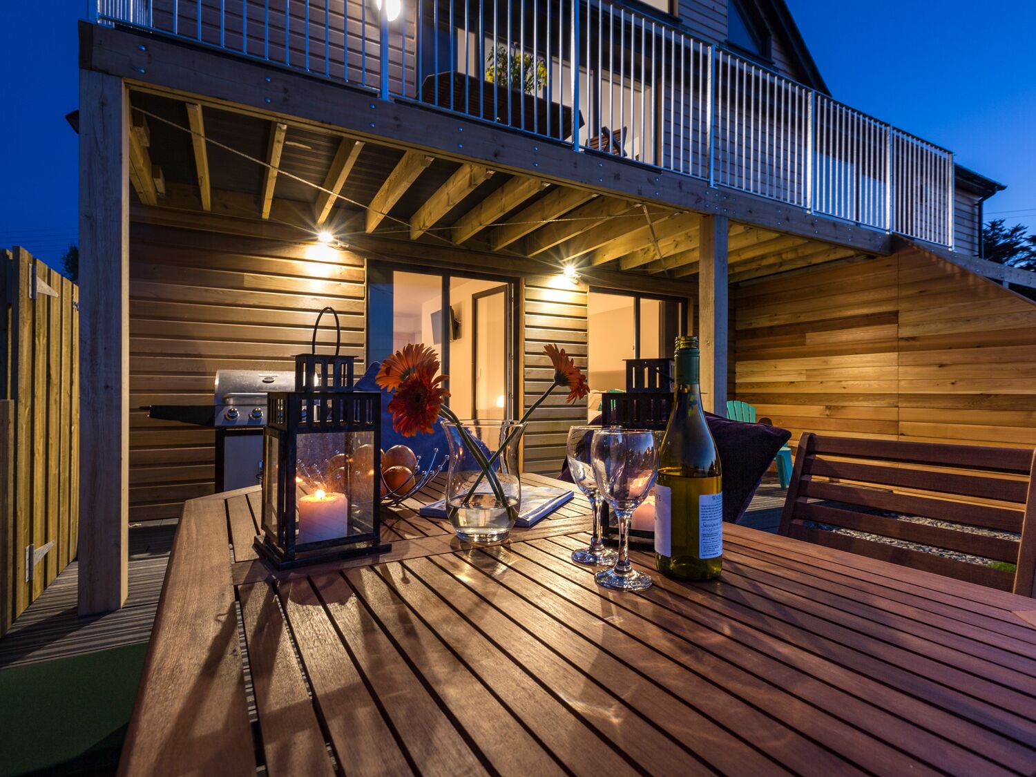 Une terrasse extérieure en bois, avec une table dressée pour les repas, un barbecue et une lanterne.