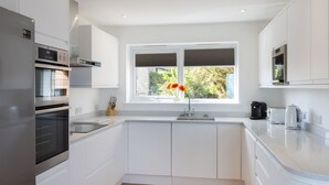 A modern kitchen with white cabinetry, a stainless steel refrigerator, a built-in oven, a double sink, and a window with a view of greenery. - Godrevy (St Ives)