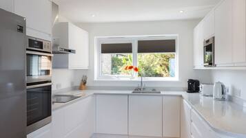 A modern kitchen with white cabinetry, a stainless steel refrigerator, a built-in oven, a double sink, and a window with a view of greenery.