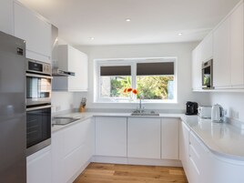 Una cocina moderna con armarios blancos, un refrigerador de acero inoxidable, horno integrado, pileta doble y ventana con vista a la vegetación.