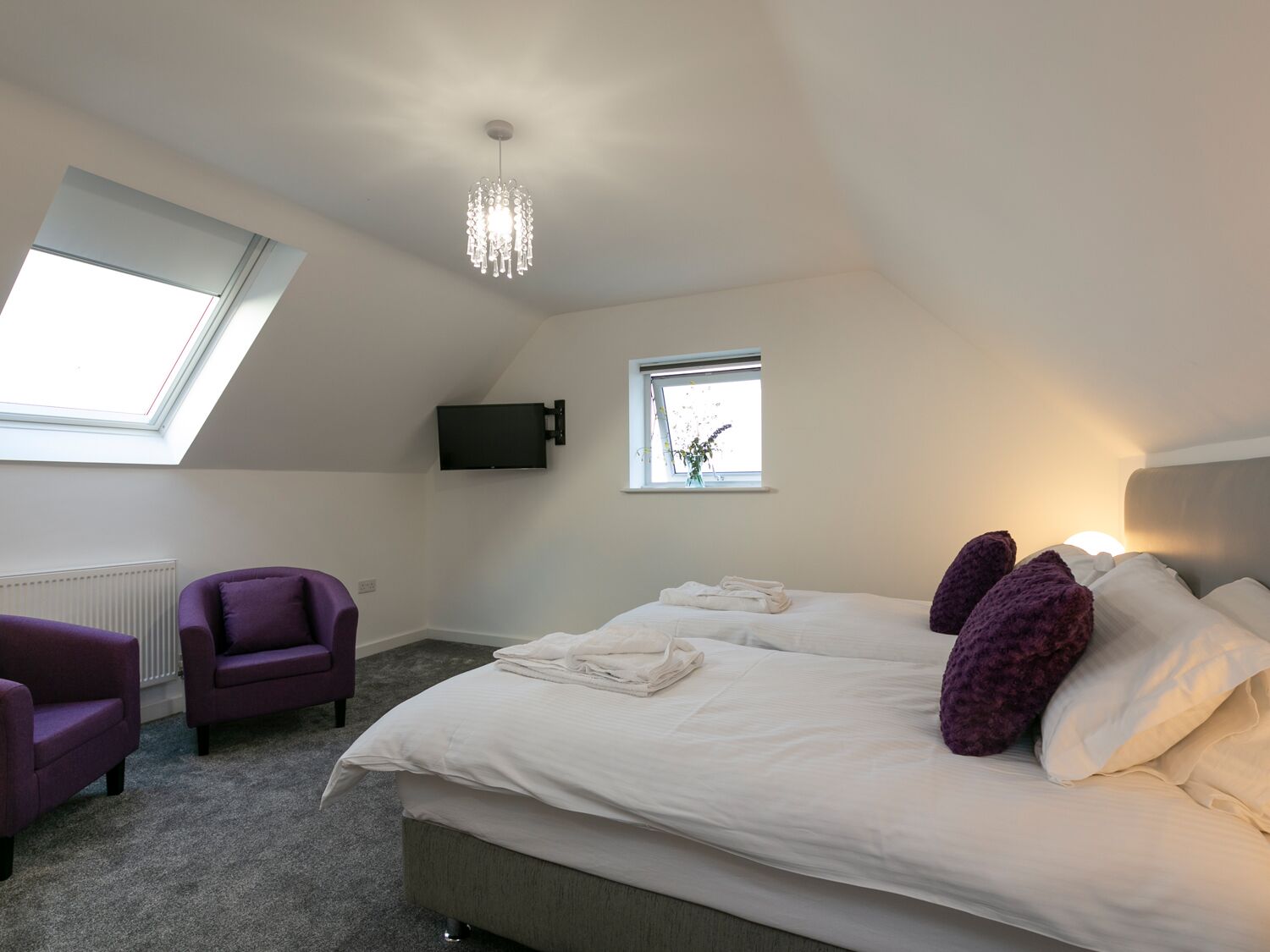 A bedroom with a large bed, two purple armchairs, a TV on the wall, and a skylight.