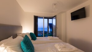 A hotel room with a large bed, a TV mounted on the wall, and a view of the sea through the sliding glass doors. - Godrevy (St Ives)