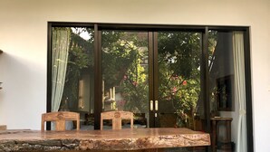 A dining area with a large wooden table and chairs, a small stool, and a glass door leading to a garden. - The Hidden Bali Inn (Ubud)