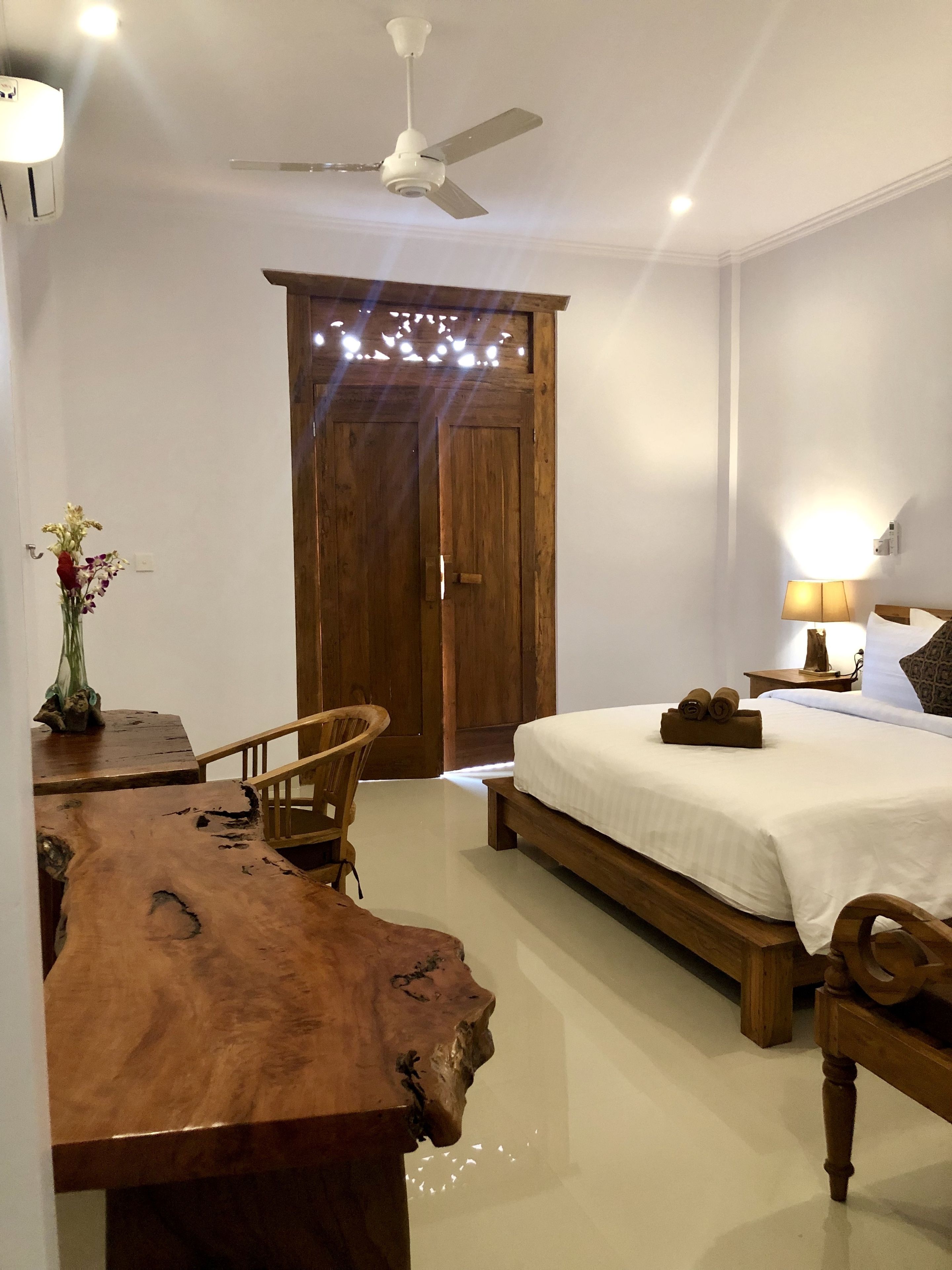 A bedroom with a wooden bed, a wooden dining table, a chair, a ceiling fan, and a wall-mounted lamp.