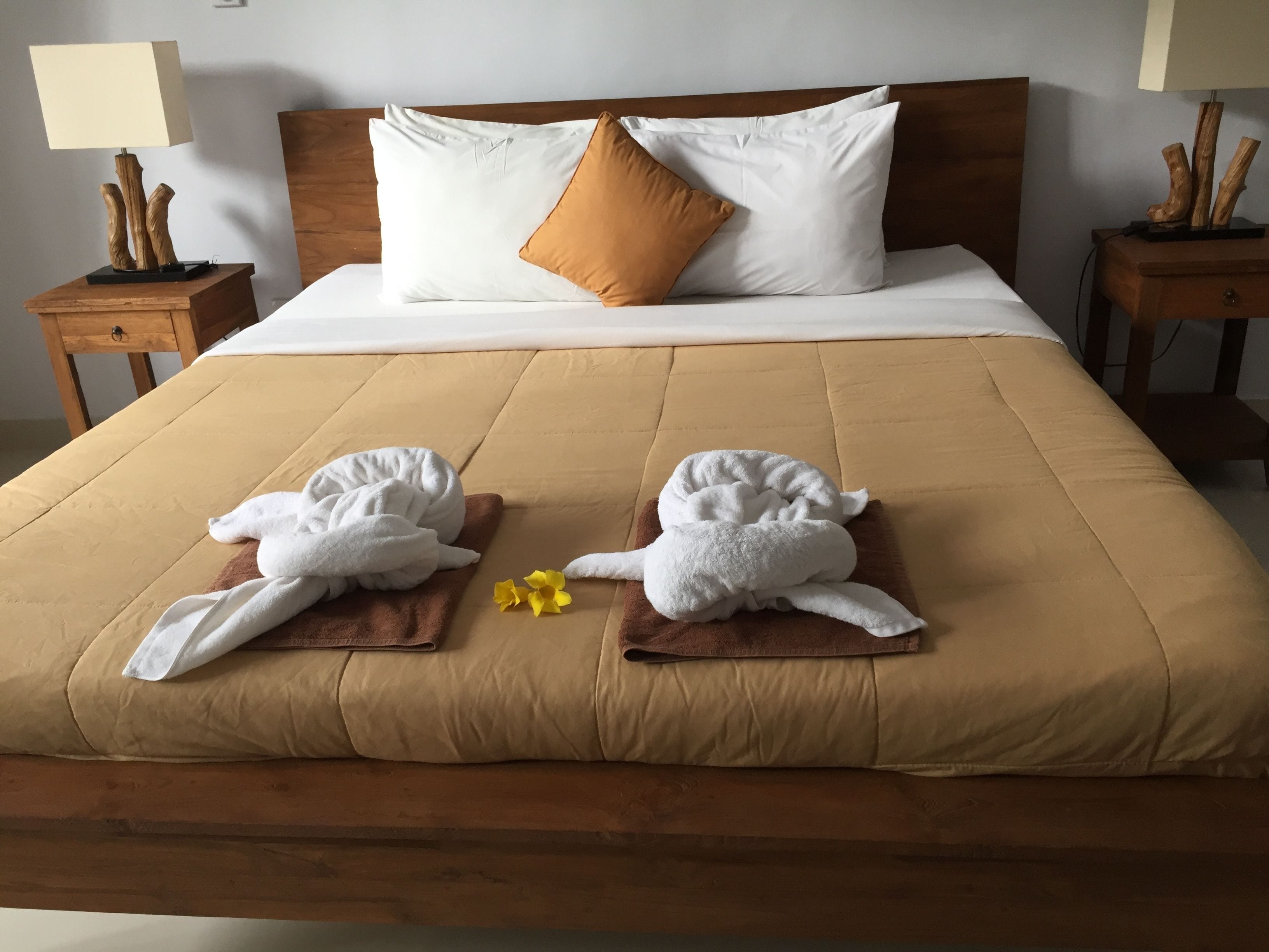 A hotel room with a large bed, wooden headboard, white and brown pillows, two wooden nightstands with lamps, and towels with yellow flowers on the bed.