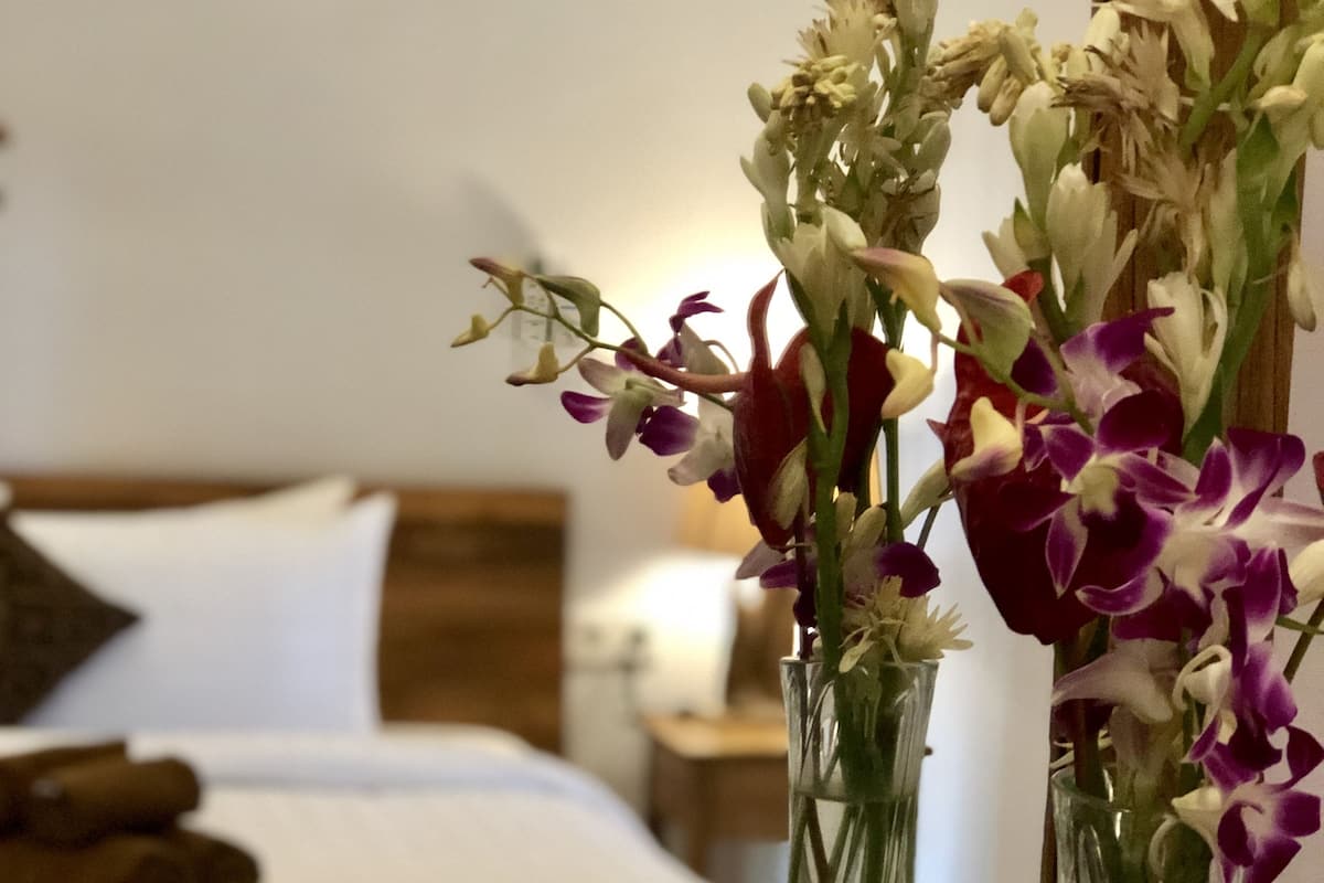 A hotel room with a bed, a wooden headboard, a nightstand, and a mirror reflecting a vase of flowers.