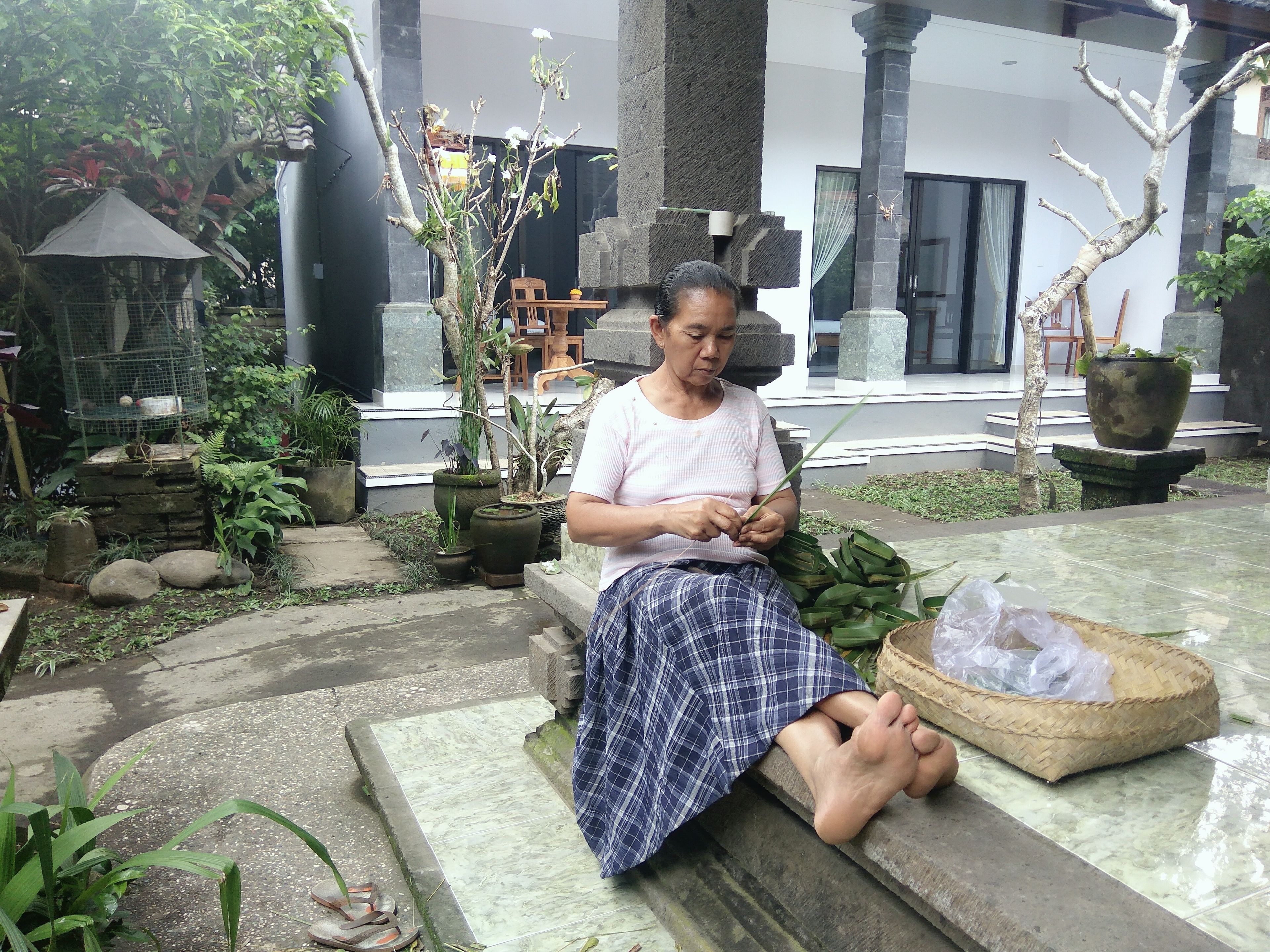 A person seated outdoors with a basket of items on a tiled surface, surrounded by potted plants and a stone structure.
