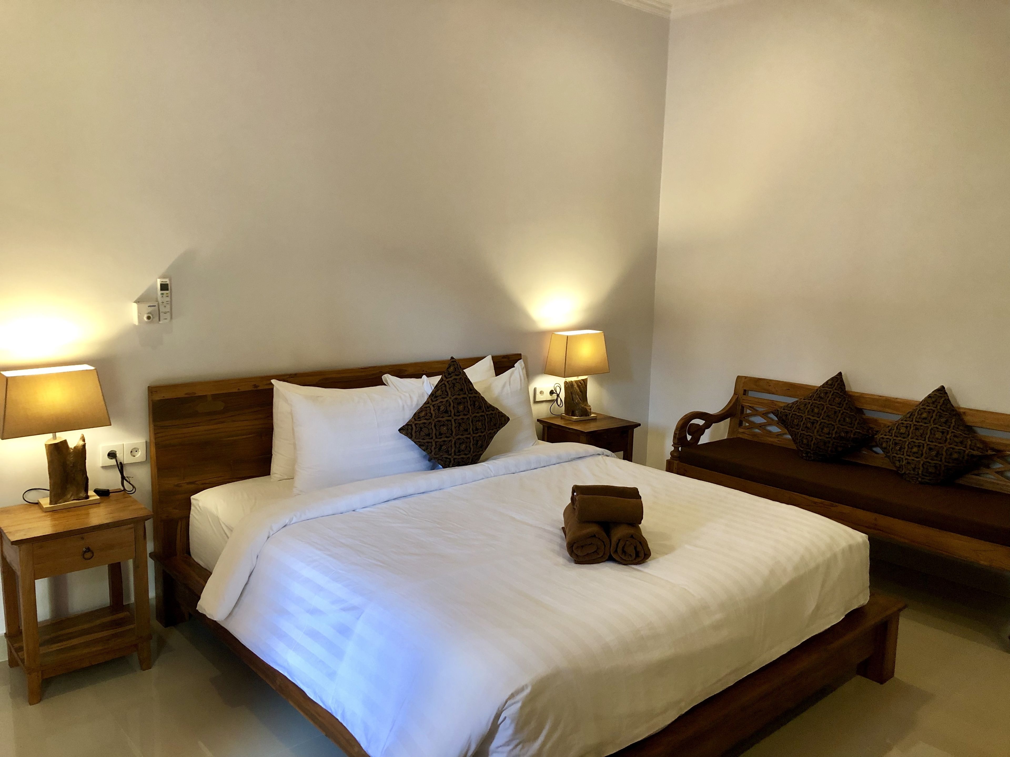 A neatly made bed with white linens and two pillows, a wooden bench with cushions, and two bedside tables with lamps.