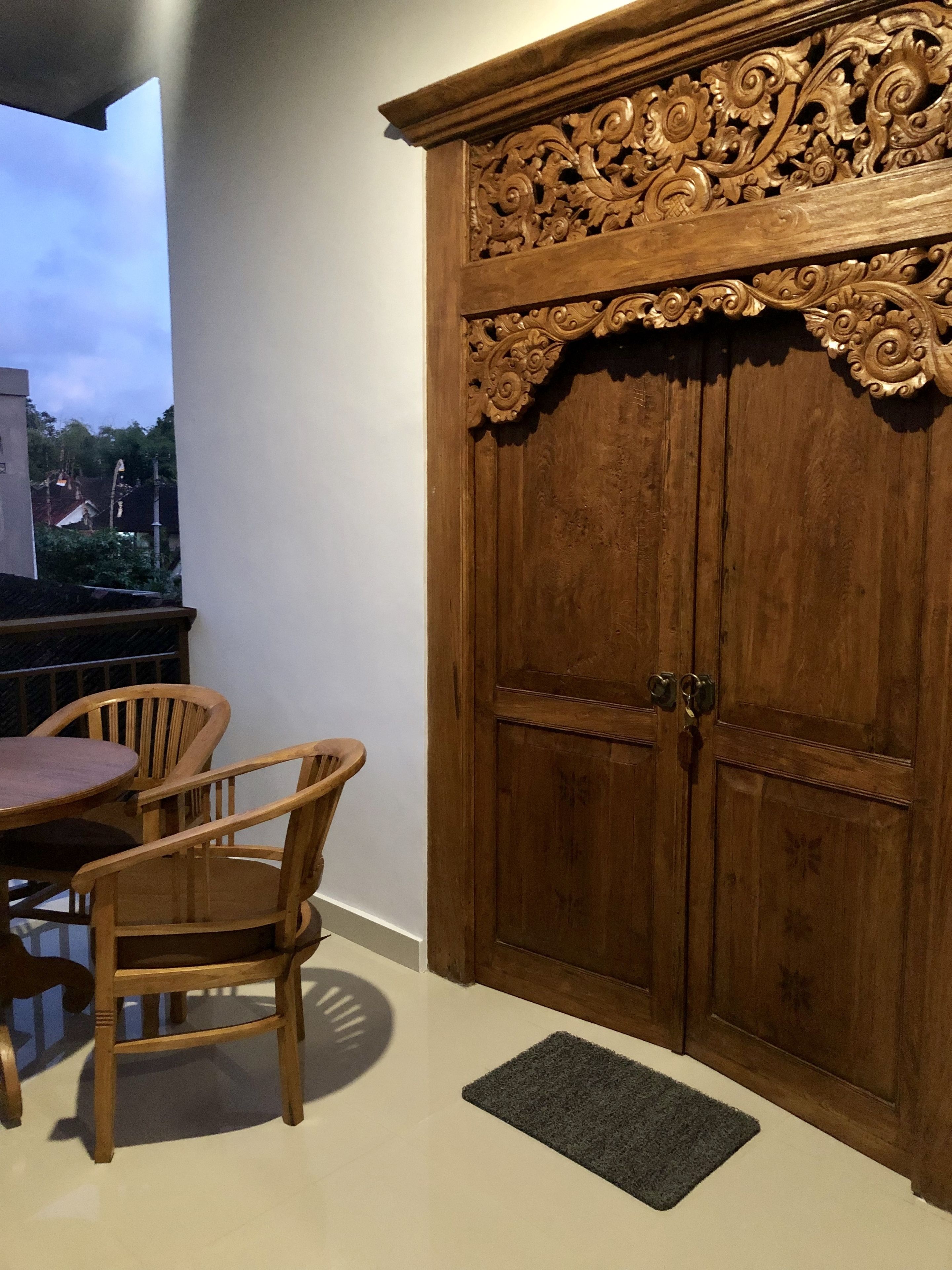 A wooden wardrobe with intricate carvings, a round wooden table with chairs, and a small rug on a tiled floor.