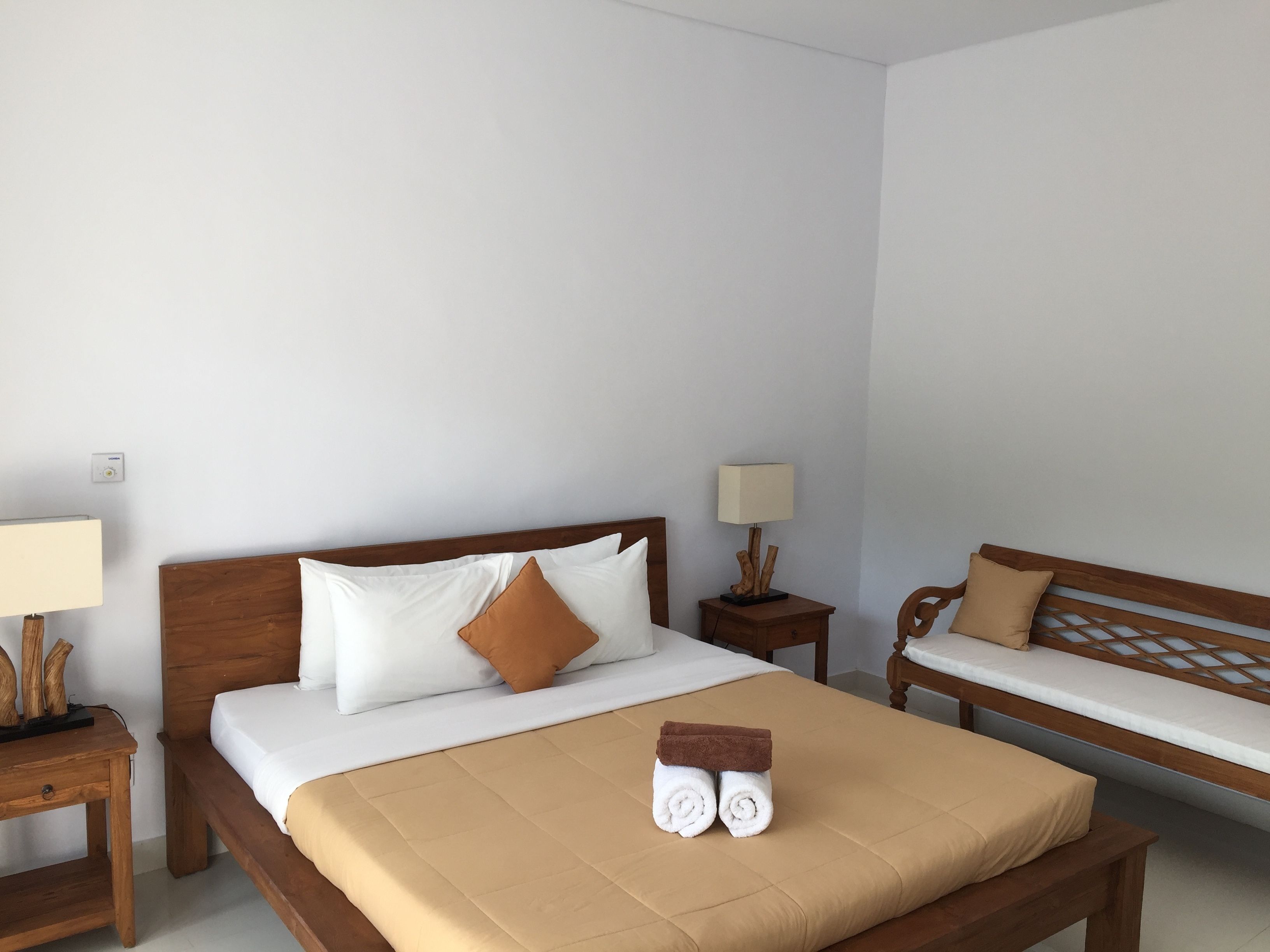 Quarto de hotel com uma cama, um banco e uma mesa de cabeceira com uma luminária.
