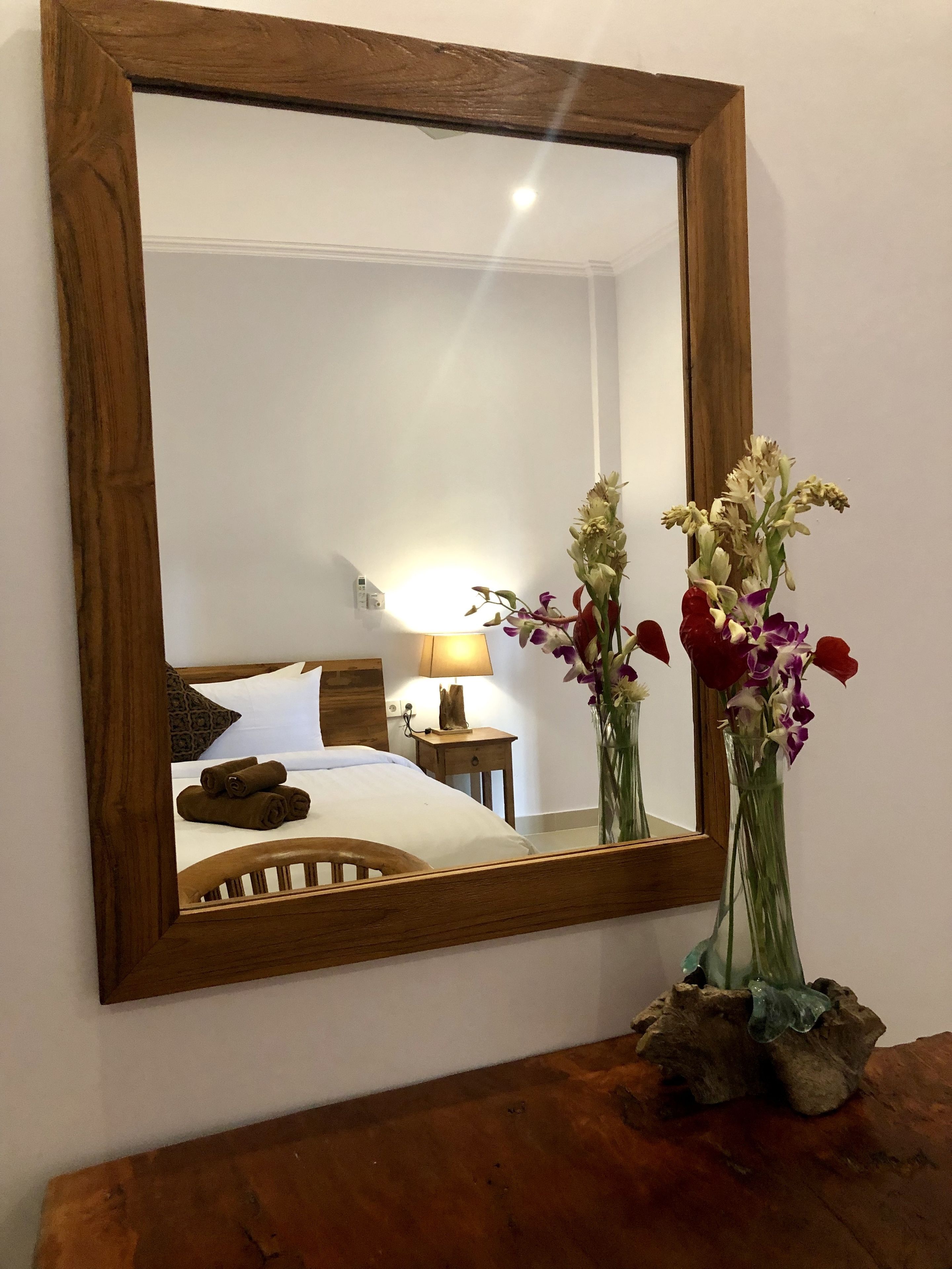 A mirror with a wooden frame reflecting a bed, pillows, a nightstand, a lamp, and a vase with flowers.