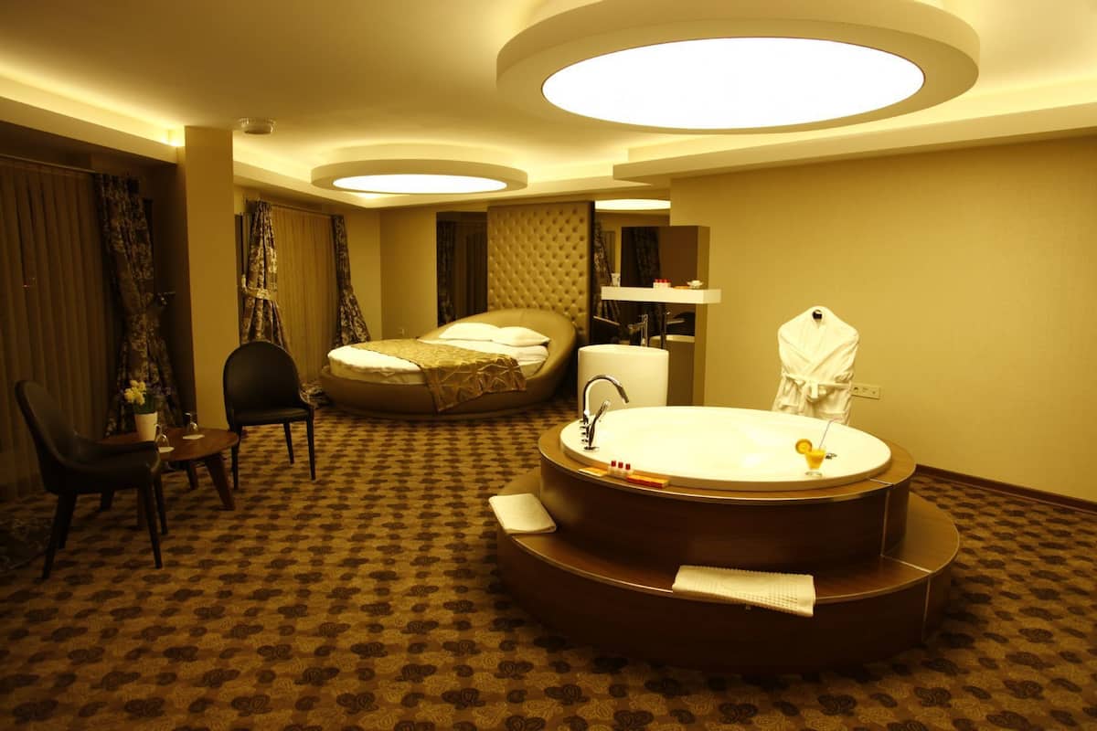 A hotel room with a large round bathtub, a bed, a chair, and a small table with a vase of flowers.