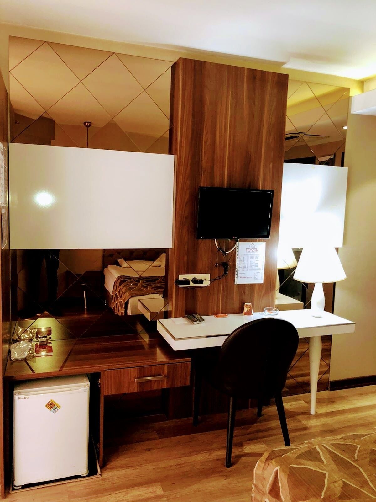 A hotel room with a wooden desk, a television mounted on the wall, a small refrigerator, a bed with a white headboard, and a bedside table with a lamp.